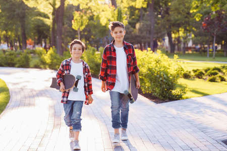 two pretty brothers of Caucasian nationality standing in the park with skateboards in hands. Dressed in jeans and plaid red shirts with white T-shirts. Smiling and enjoying a warm summer evening.の写真素材