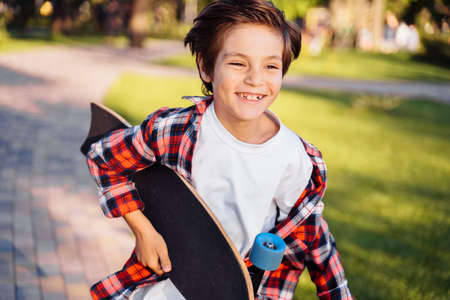 Happy young guy running along the alley on a sunny summer evening. Holding his skateboard in hands. Actively spending summer holidays. Dressed in a plaid shirt and white T-shirt.の写真素材