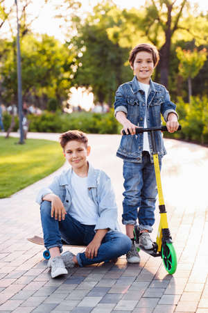 Active children's entertainment in the summer park. Two cute brothers having fun in the park on a summer evening. Riding on a skateboard and scooter. Dressed in denim suits and white T-shirts.の写真素材