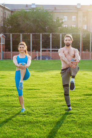 Attractive young couple man and woman 25 years old are doing exercises on a sunny summer morning on grass sports stadium. Joyfully and with a smile making slopes, stretching muscles, raising legs.の写真素材