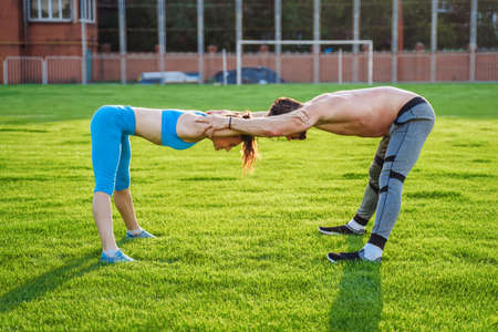 Attractive young couple man and woman 25 years old are doing exercises on a sunny summer morning on grass sports stadium. Joyfully and with a smile making slopes, stretching muscles, raising legs.の写真素材