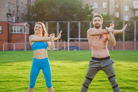 Attractive young couple man and woman 25 years old are doing exercises on a sunny summer morning on grass sports stadium. Joyfully and with a smile making slopes, stretching muscles, raising legs.の写真素材