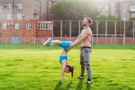 Man standing on his hands upside down and woman holding him. Strong, muscular extreme sportsmen. Young athletes of Caucasian nationality standing on the football field and doing physical exercises.の写真素材