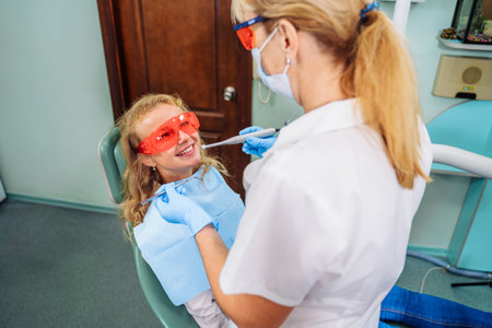 Charming girl patient sitting at the reception in the dental chair. She is wearing red safety glasses that protect against harmful radiation. Dental procedures in a modern, equipped office.の写真素材