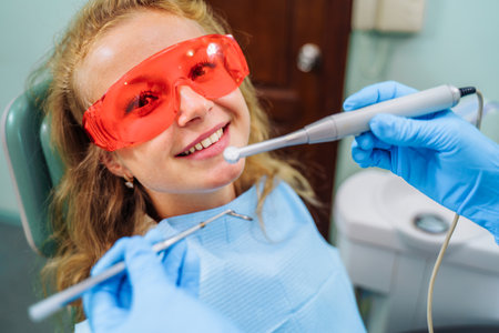 Charming girl patient sitting at the reception in the dental chair. She is wearing red safety glasses that protect against harmful radiation. Dental procedures in a modern, equipped office.の写真素材