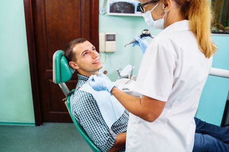 A young, attractive European-looking guy came to the dentist. Sits in a dental chair. The dentist bent over him and prepares to treat problem teeth. Happy patient and dentist concept.の写真素材