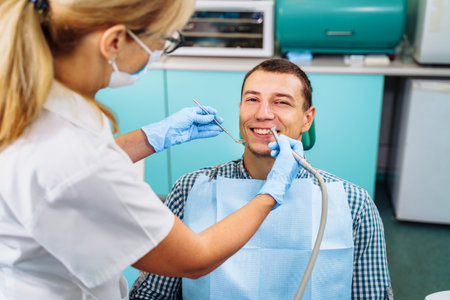 A young, attractive European-looking guy came to the dentist. Sits in a dental chair. The dentist bent over him and prepares to treat problem teeth. Happy patient and dentist concept.の写真素材