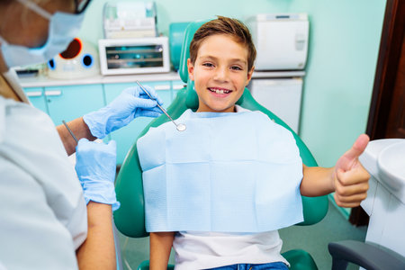 Small, young patient boy is happy with a visit to the dentist. Concept of painless dental treatment. Beautiful, wide smile of a child sitting in a dental chair in a doctor's office. Thumbs up.の写真素材