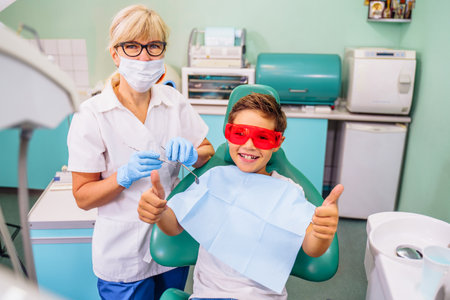 Small, young patient boy is happy with a visit to the dentist. Concept of painless dental treatment. Beautiful, wide smile of a child sitting in a dental chair in a doctor's office. Thumbs up.の写真素材