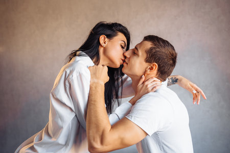 Attractive, young couple of lovers having sex at home in the bed in bright room. Girl kissing her boyfriend oddly, gently pulling back his lower lip. Celebrating Valentine's Day - February 14.の写真素材