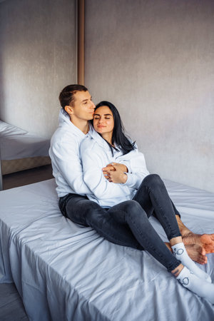 Beautiful young couple in the same white hoodie and black pants lie on the bed hugging. Dreaming of happy future together. Enjoying each other's company. Man gently hugs his beloved from back.の写真素材