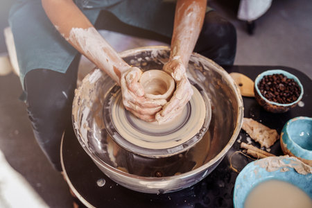 Close-up of the hands of a woman ceramist working with a potter's wheel in a cozy, bright workshop. A young experienced woman skillfully makes a mug, vase or plate. Creative people, pottery workshop.の写真素材