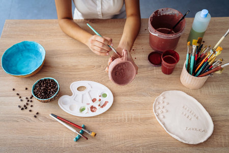 Female hands over workshop table with clay dish, bowl, paintbrushes and paint samples, coloring mug. Overhead shot of artistic tools for pottery making.の写真素材