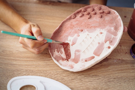 Close-up of hands. Selective focus. A young woman artist painting her clay product, a plate, in brown. Preparing for final burning. Brush on a colorless plate to make it bright and colorful later.の写真素材