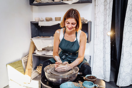 Pottery. A young woman making vase, mug, plate, pot in her own working place. Sitting at the table near the potter's wheel, creating the shape of the future product with his hands.の写真素材