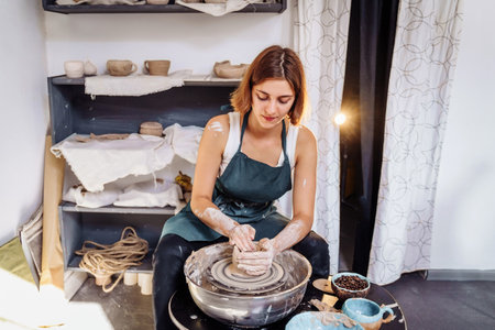 With professional hand movements, a female potter prepares a piece of clay on a potter's wheel, kneads the clay and rolls it out to make a bowl, vase, jug, plate. Pottery studio. Young professional.の写真素材