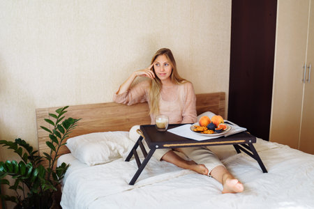 Efficient young millennial attractive woman sitting on a bed in the morning. Hand holding a cup of coffee and eating cookies. Coffee morning concept. Pleasant time during calm morning.の写真素材