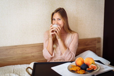 Good morning. Attractive girl drinking morning coffee in bed. Sitting on snow-white bed in the bedroom, in front of her stands for breakfast with various bright fruits and pastries. Pleasant and calm.の写真素材