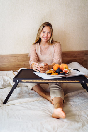 Efficient young millennial attractive woman sitting on a bed in the morning. Hand holding a cup of coffee and eating cookies. Coffee morning concept. Pleasant time during calm morning.の写真素材