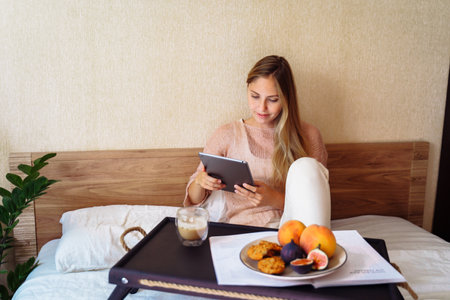 Beautiful young caucasian woman sitting on bed with sleepy look in morning. Working on bed remotely, working from home, busy lifestyle concept. Surfing internet, dressed in casual domestic clothes.の写真素材