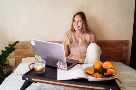Distance learning, working, online education. Happy pretty woman working remotely from home on bed instead of office. Using computer, laptop and drinking coffee. Smiling and thinking.の写真素材