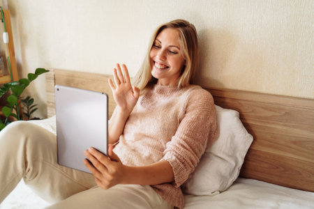 Young, pretty, attractive, charming girl blonde of European appearance, is smiling widely on tablet with friends and relatives. Using video communication, friendly waving hand, looking at the screen.の写真素材