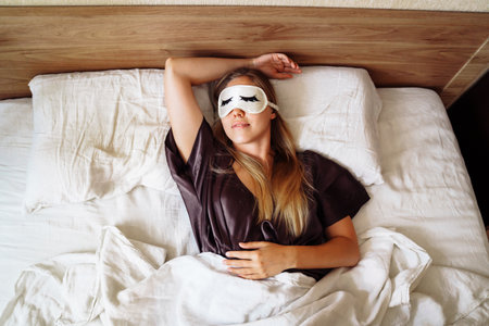 Pretty girl in night robe sleepping sweetly in snow-white bed in her room. View from the top, above. Wearing eyelash-patterned night goggles. Cozy covered with a blanket. Sweet morning dream at dawn.の写真素材