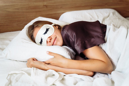 A young beautiful woman is sleeping in her bed with a sleep mask against a bright, annoying light. Relaxing morning rest. A comfortable bed with white bedding in a bright bedroom. Hugging a pillow.の写真素材