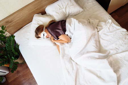 Pretty girl in night robe sleepping sweetly in snow-white bed in her room. View from the top, above. Wearing eyelash-patterned night goggles. Cozy covered with a blanket. Sweet morning dream at dawn.の写真素材