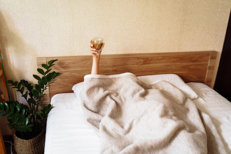A sleepy young woman lies in a snow-white bed. Covered with beige blanket over her head. Raised her hand with a cup of coffee. It is impossible to wake up without a cup of invigorating coffee.の写真素材
