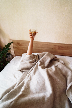 A sleepy young woman lies in a snow-white bed. Covered with beige blanket over her head. Raised her hand with a cup of coffee. It is impossible to wake up without a cup of invigorating coffee.の写真素材