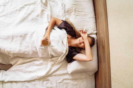 Young woman lies in a beautiful, white bed. Almost woke up. Early morning. Covers face with hands, shading eyes from the light. Difficulty getting up for owl-type people. Laughs, smiles to new day.の写真素材