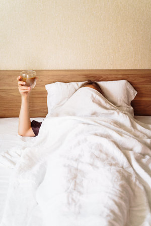 A sleepy young woman lies in a snow-white bed. Covered with beige blanket over her head. Raised her hand with a cup of coffee. A funny shot about the difficulty of waking up in the morning.の写真素材