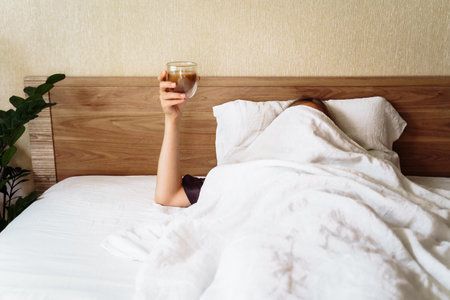A sleepy young woman lies in a snow-white bed. Covered with beige blanket over her head. Raised her hand with a cup of coffee. A funny shot about the difficulty of waking up in the morning.の写真素材