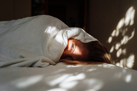 Beautiful, young blonde lies on the bed in the rays of soft, morning sun. Buried under the covers, showing out small part of her face. Wakes up slowly. Difficulty getting up, let me sleep some more.の写真素材