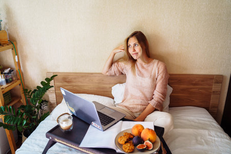 Young woman using laptop in a cozy home atmosphere on the bed with a cup of coffee and delicious fruits with pastries. Freelancer cam choose where to work. Earnings at home. Internet gives freedom.の写真素材