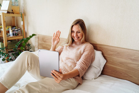 Young, pretty, attractive, charming girl blonde of European appearance, is smiling widely on tablet with friends and relatives. Using video communication, friendly waving hand, looking at the screen.の写真素材