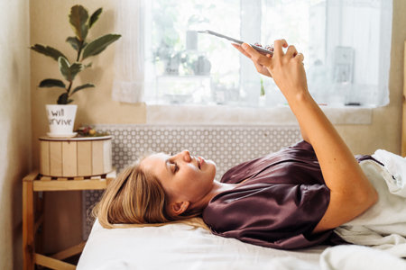 Side view. The attractive, sweet blonde lies on her back on a bed in her own bedroom. Holding a tablet in her hands and reading, flipping through morning news. Cozy bright room in rays of summer sun.の写真素材