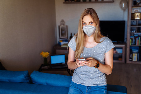 Beautiful girl reading news on the phone about the  epidemic and calling her friends. She remains isolated at home alone and wearing protective respirator while working remotely.の写真素材