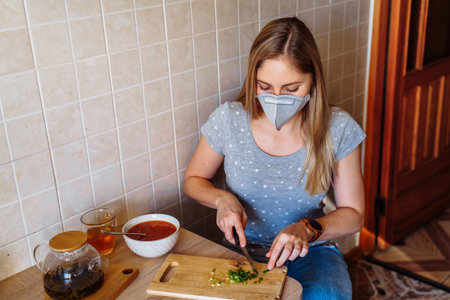 Attractive blonde is sitting in the kitchen near the table with food. She is isolated at home during quarantine due to the  epidemic, wearing a protective mask.Looking to plate with soup.の写真素材