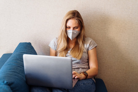 Attractive blonde girl wearing a protective mask works hard and responsibly with a laptop remotely at home. Isolated due to the worldの写真素材