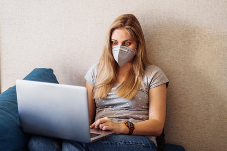 Attractive blonde girl wearing a protective mask works hard and responsibly with a laptop remotely at home. Isolated due to the worldの写真素材