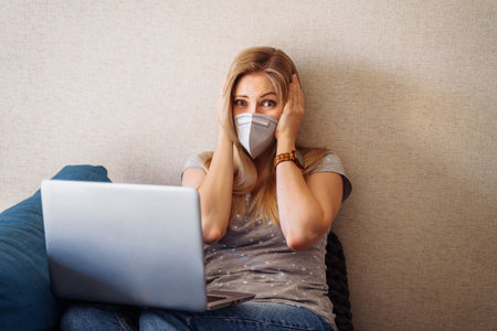 Anxious caucasian girl clutching her head with hands in alarm and looking to her laptop at the horrific numbers of statistics on sick and dead during the  pandemic. Wearing face mask.の写真素材