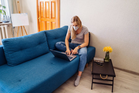 Attractive blonde girl wearing a protective mask works hard and responsibly with a laptop remotely at home. Isolated due to the world epidemic during the quarantine periodの写真素材