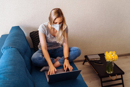 Attractive blonde girl wearing a protective mask works hard and responsibly with a laptop remotely at home. Isolated due to the world   epidemic during the quarantine periodの写真素材