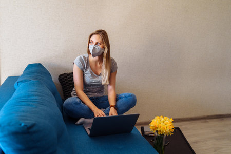Attractive blonde girl wearing a protective mask works hard and responsibly with a laptop remotely at home. Isolated due to the world   epidemic during the quarantine periodの写真素材