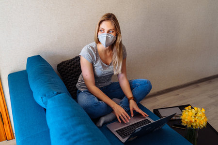 Attractive blonde girl wearing a protective mask works hard and responsibly with a laptop remotely at home. Isolated due to the world  epidemic during the quarantine periodの写真素材