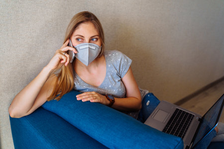 Casual girl in a protective face mask is phoning on labor issues. Isolated at home due to epidemic and high risk of getting sick and die.の写真素材