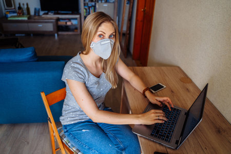 Beautiful girl sitting at home and looking to the camera, wearing protective face mask. Isolated at home due to pandemic. Remote work for health protectionの写真素材