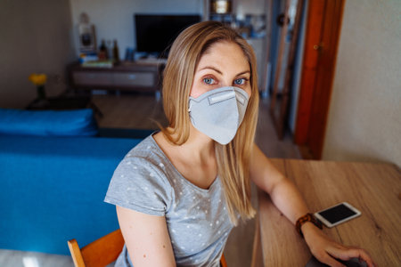 Beautiful girl sitting at home and looking to the camera, wearing protective face mask. Isolated at home due to pandemic. Remote work for health protectionの写真素材